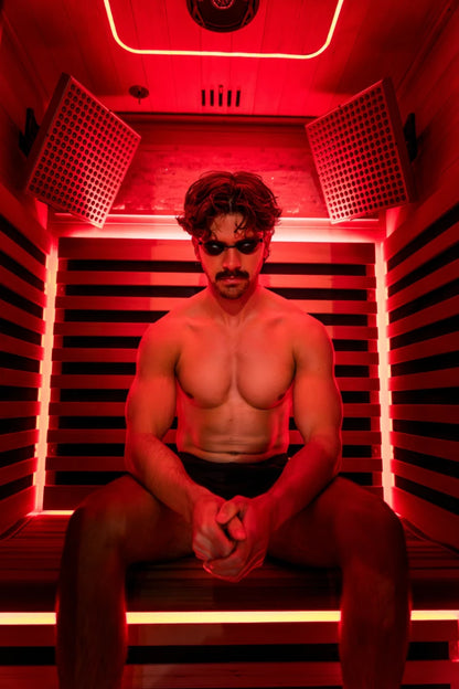 Man sitting inside a red light therapy sauna chamber with dual heat panels on either side, neon accents around the frame, and ventilation grilles visible at the top.