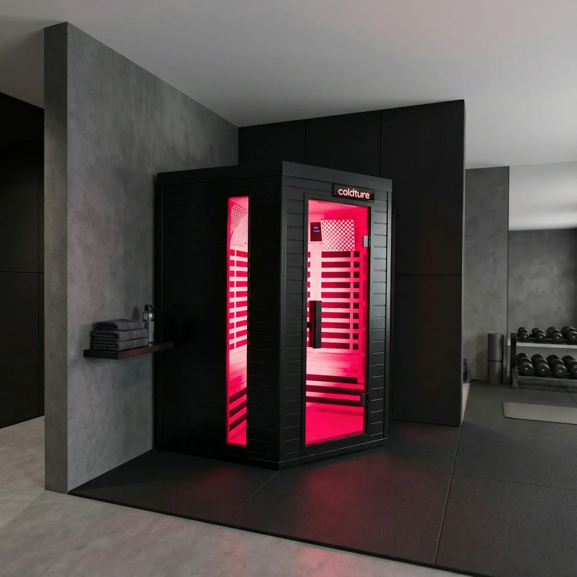 Black infrared sauna pod with bright pink LED light interior, glass door panel, and "Coldture" branding, positioned in a modern gym setting with concrete walls and dumbbells visible nearby.