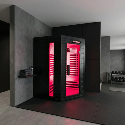 Black infrared sauna pod with bright pink LED light interior, glass door panel, and "Coldture" branding, positioned in a modern gym setting with concrete walls and dumbbells visible nearby.