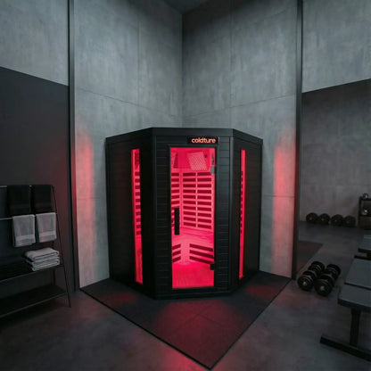 Black corner infrared sauna with bright red interior lighting and horizontal bench seating, installed in a modern gym with concrete walls and dumbbells nearby.