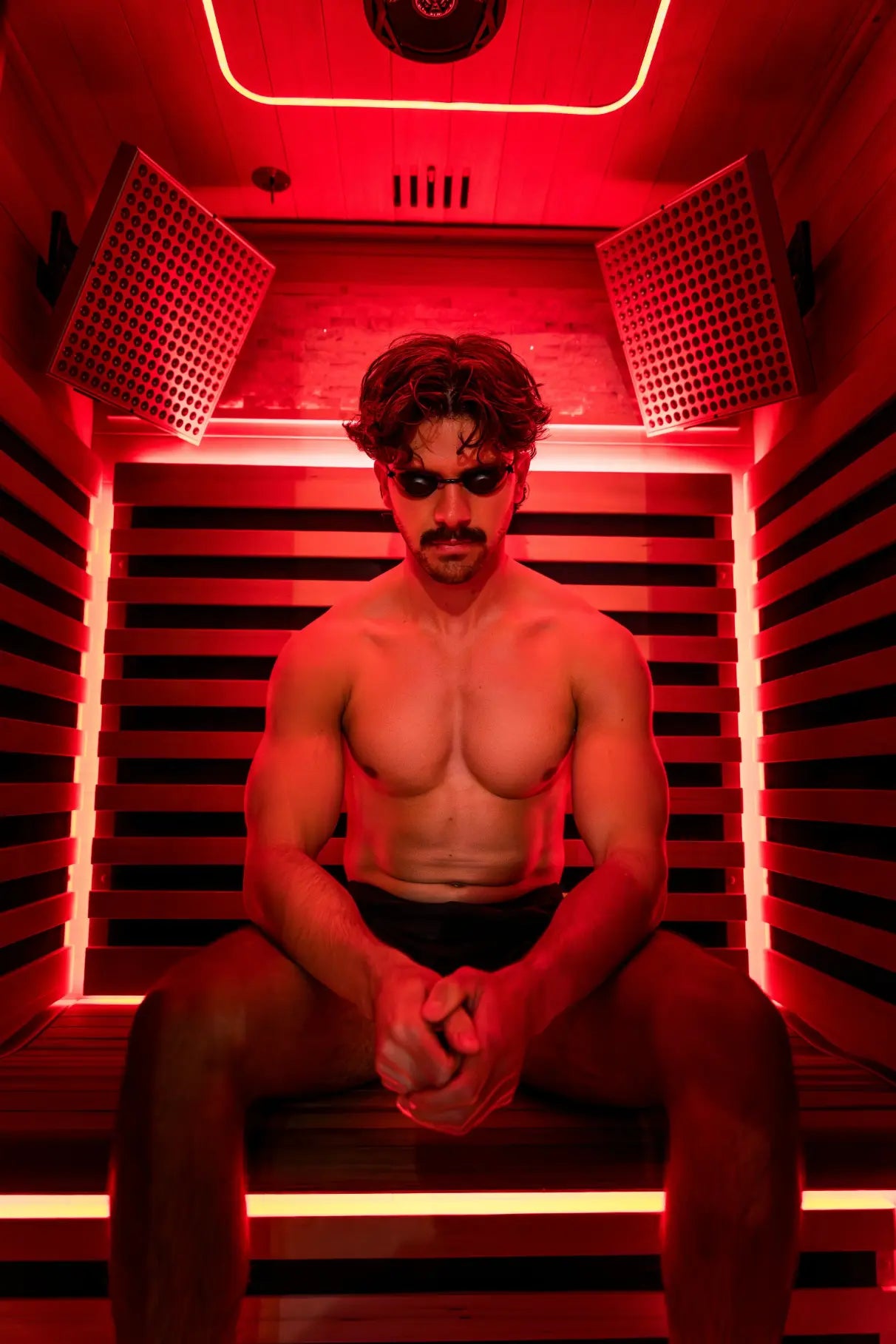 Man seated inside a red-light infrared sauna pod with gridded panel lights on either side and neon tubing around the interior frame.