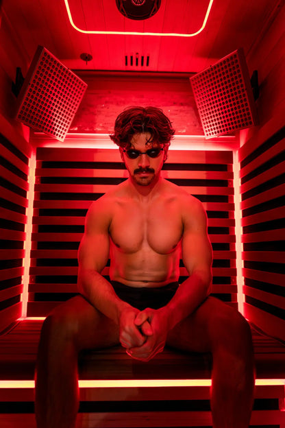 Man seated inside a red-light infrared sauna pod with gridded panel lights on either side and neon tubing around the interior frame.