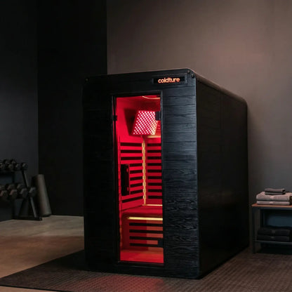 Black infrared sauna cabinet with red interior lighting, glass door panel, and "coldture" branding visible on top. Wooden bench seating and heat lamp visible inside.