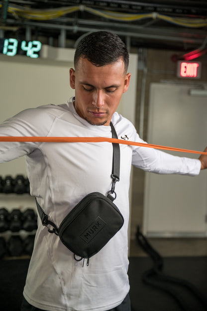 Man stretching with orange resistance band in gym, wearing white shirt and black crossbody bag.