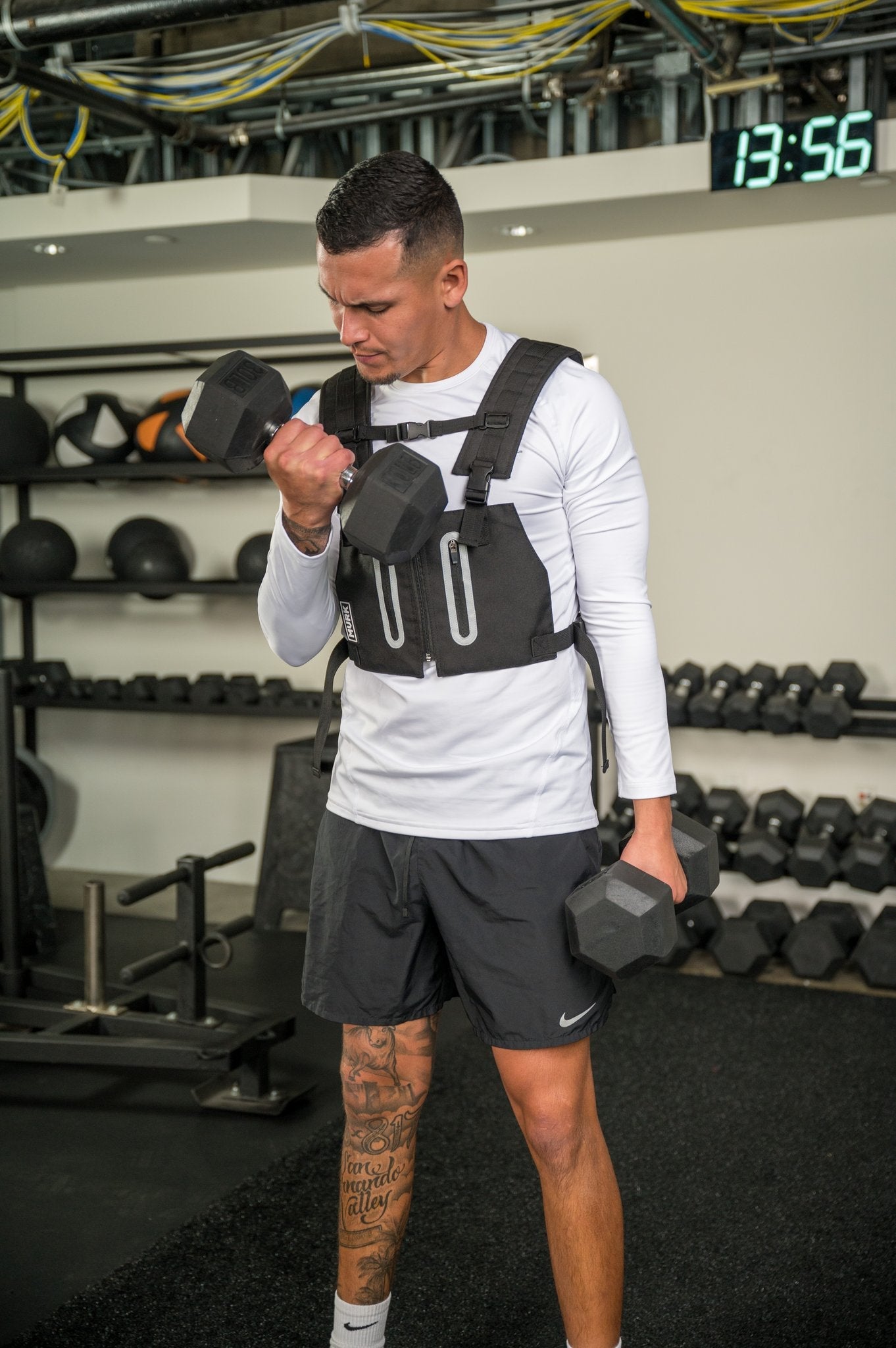 Man in gym wearing a weighted vest and lifting dumbbells.
