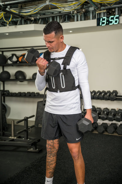 Man in gym wearing a weighted vest and lifting dumbbells.