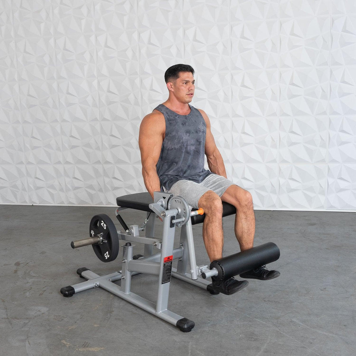 Man wearing a dark gray tank top sits on a leg extension machine in a gym.
