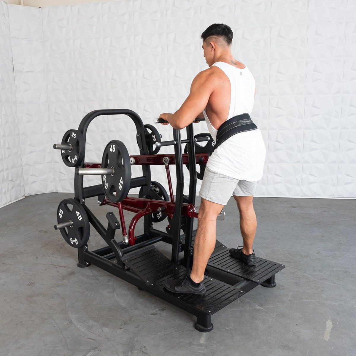 Man in white tank top using weight-plate machine with belt.