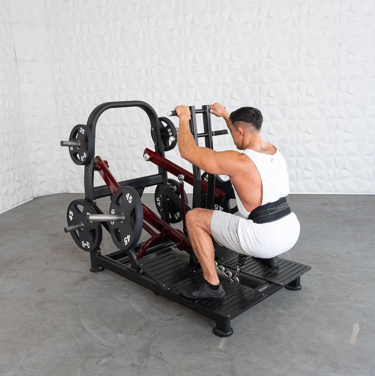 Man in white tank top uses plate-loaded leg extension machine at gym.