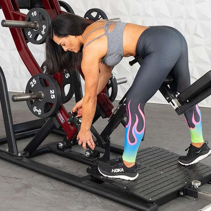 Woman adjusting lever on fitness machine, wearing colorful flame leggings and Hoka shoes in gym setting.