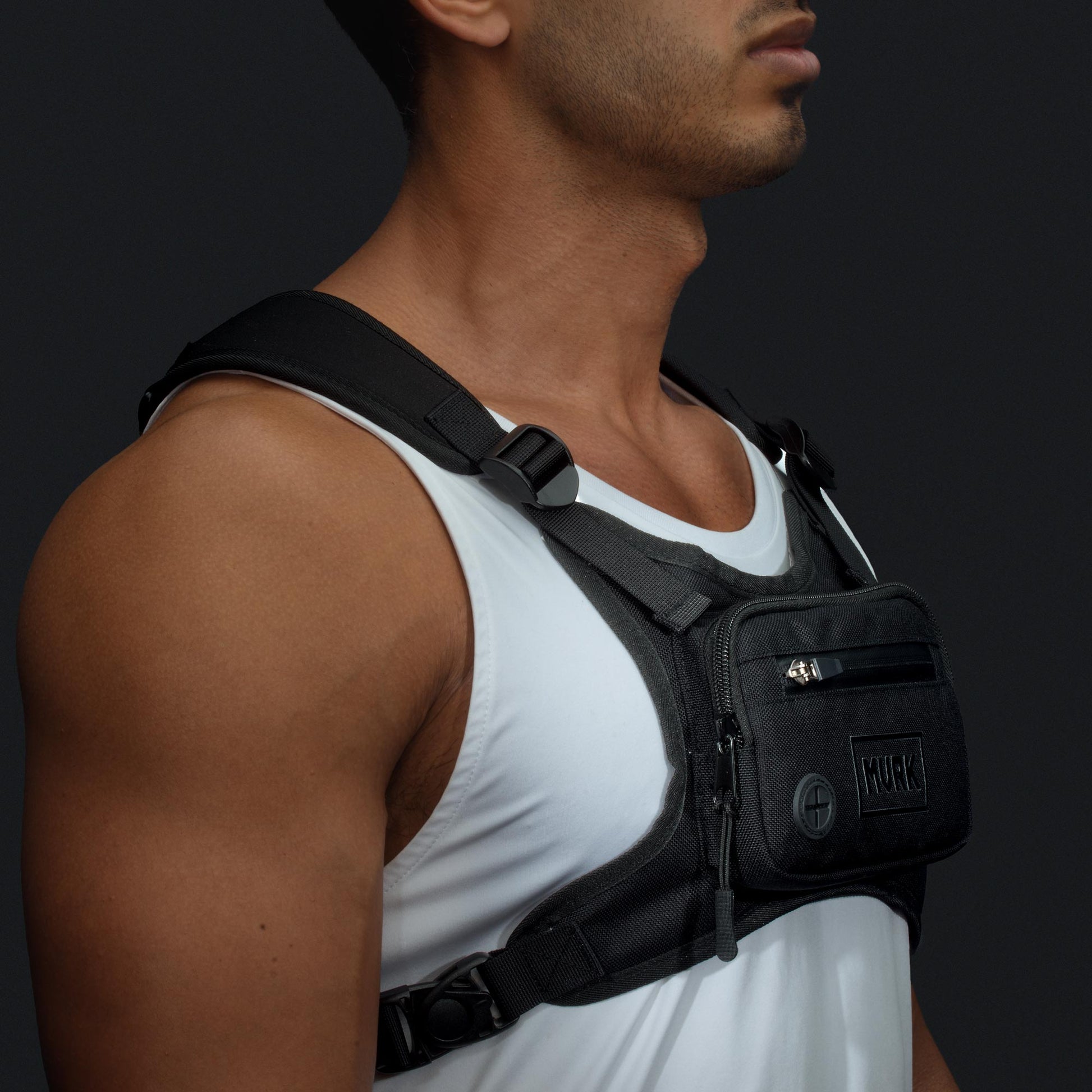 Man in a white tank top wearing a black chest rig with small pockets.