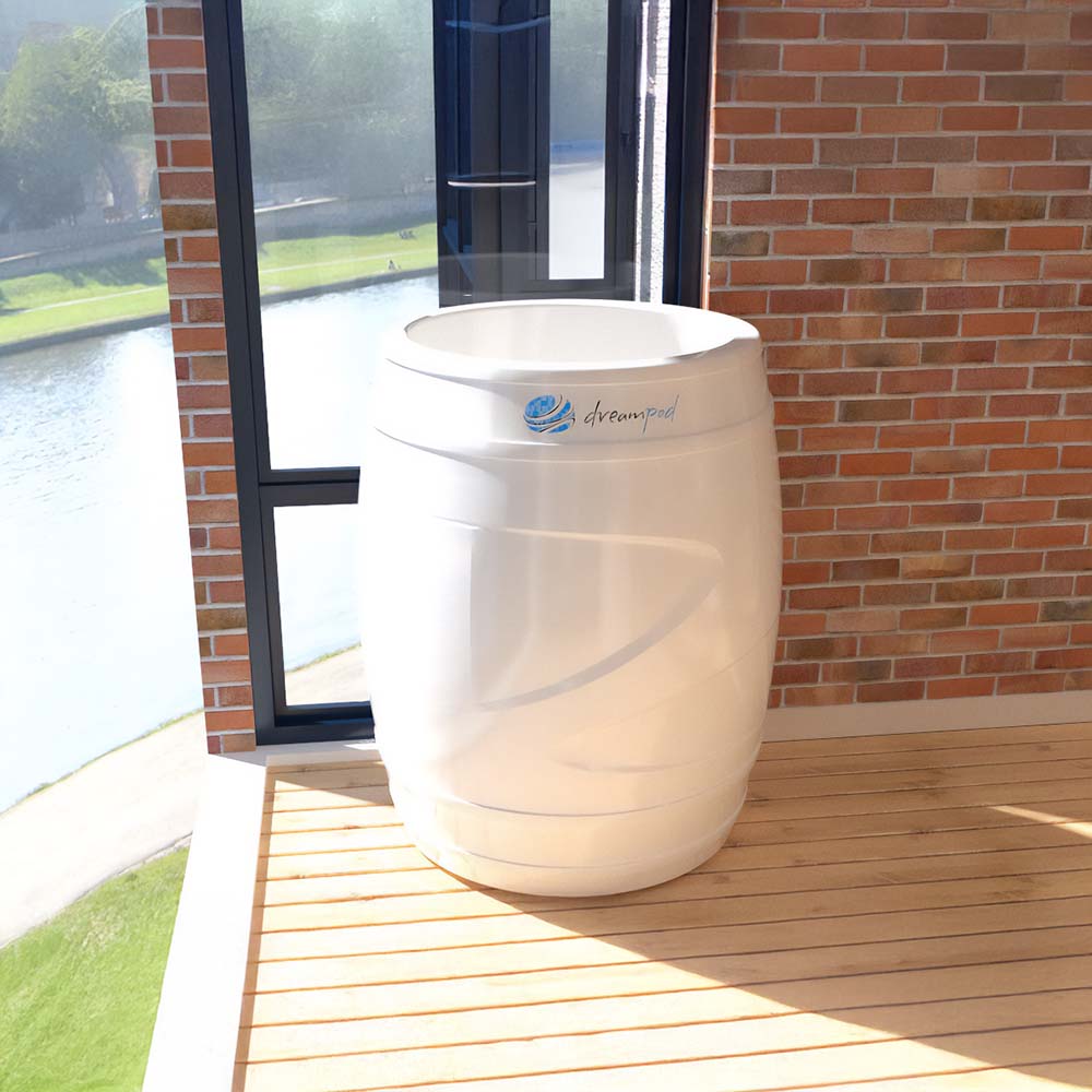 A Dreampod Cold Plunge Barrel displayed in a bright room with large windows, overlooking a scenic view of a river and green landscape. The barrel is white and positioned on a wooden deck.