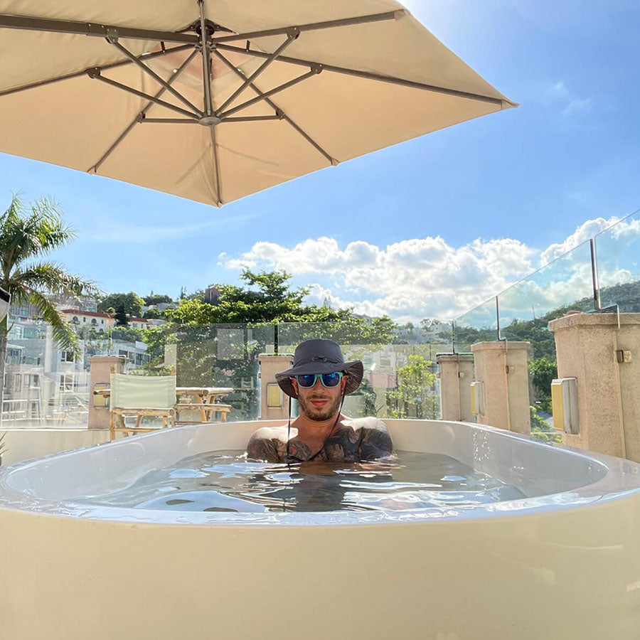 A person relaxing in a Dreampod Commercial Cold Plunge tub under a large umbrella, with a scenic view of greenery and blue sky in the background.