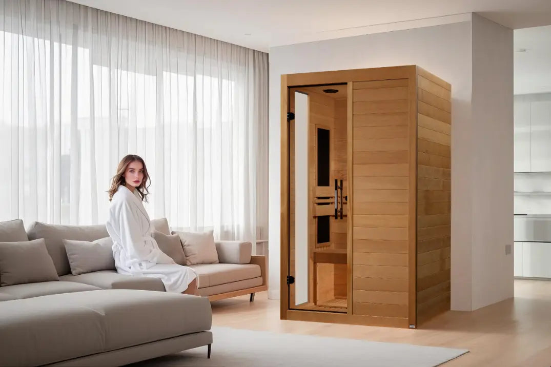 Woman in robe sits on sofa beside a two-person infrared sauna in a bright living room.