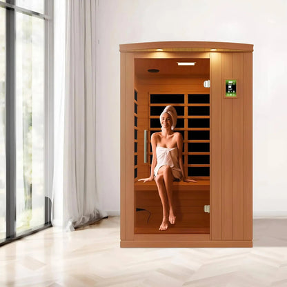 A woman sitting inside a Dynamic Venice 2 Person Far Infrared Sauna, wearing a towel and a head wrap, with a bright, modern interior setting featuring large windows.