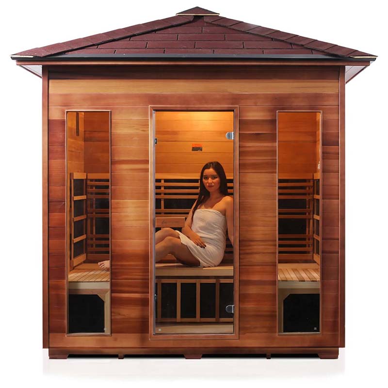 Woman relaxing in a red cedar outdoor sauna with glass doors, enjoying infrared heat therapy.
