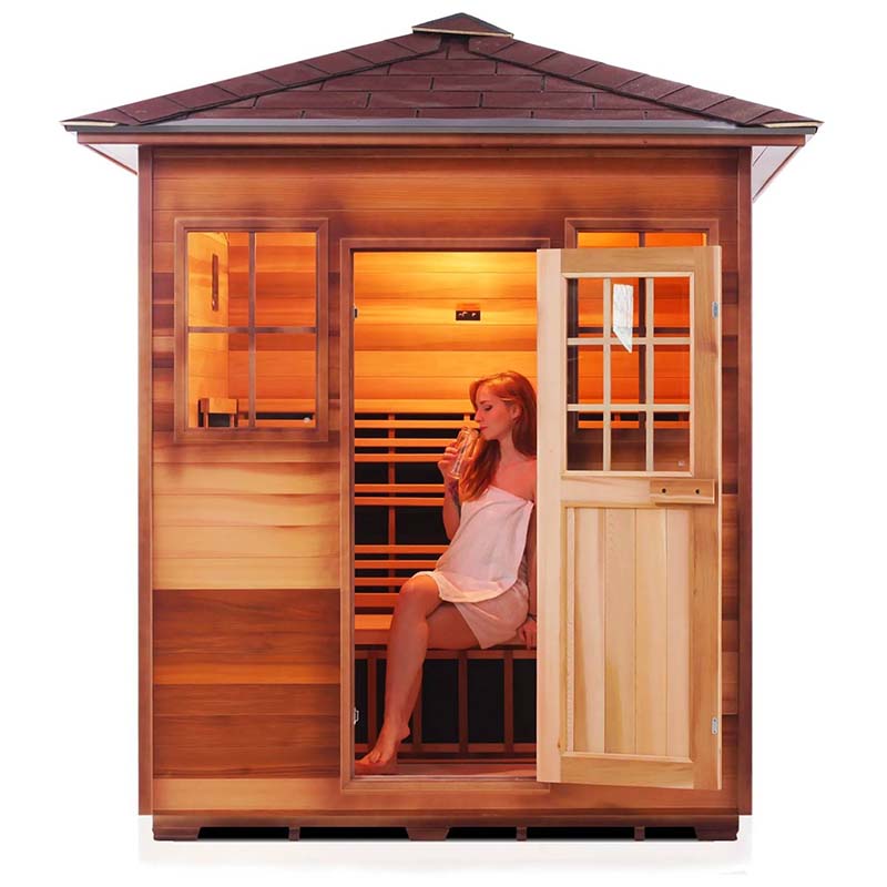 Woman relaxing in a wooden sauna, drinking water, with open door showing infrared heating panels.