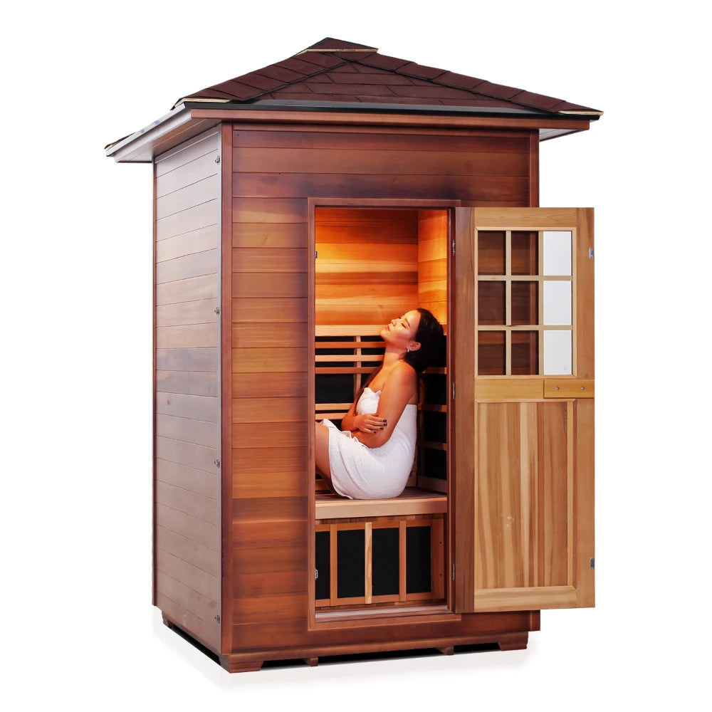 A woman sitting inside a wooden sauna, wrapped in a towel, with the door open and warm light illuminating the interior.