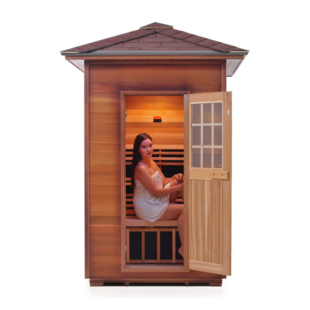 A woman sitting inside a wooden sauna with an open door, wearing a white towel, and looking relaxed. The sauna features a sloped roof and warm wooden interior.