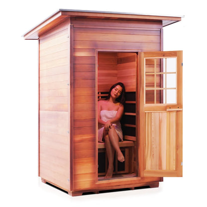 A woman sitting inside a wooden sauna, wrapped in a towel, with a serene expression. The sauna has an open door, showcasing its wooden interior.
