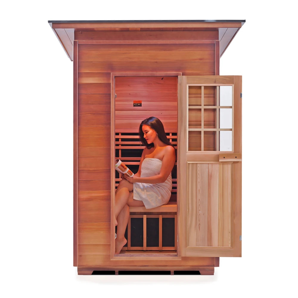 A woman wrapped in a towel sits inside a wooden sauna, reading a book. The sauna has a wooden exterior and an open door, revealing a warm interior with wooden benches.