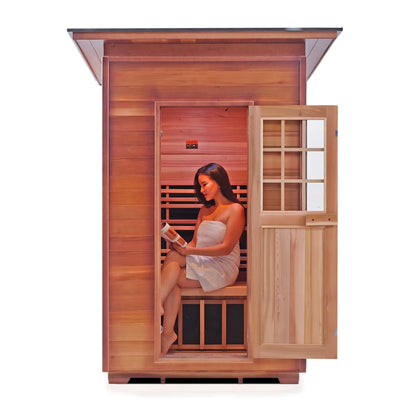 A woman wrapped in a towel sits inside a wooden sauna, reading a book. The sauna has a wooden exterior and an open door, revealing a warm interior with wooden benches.