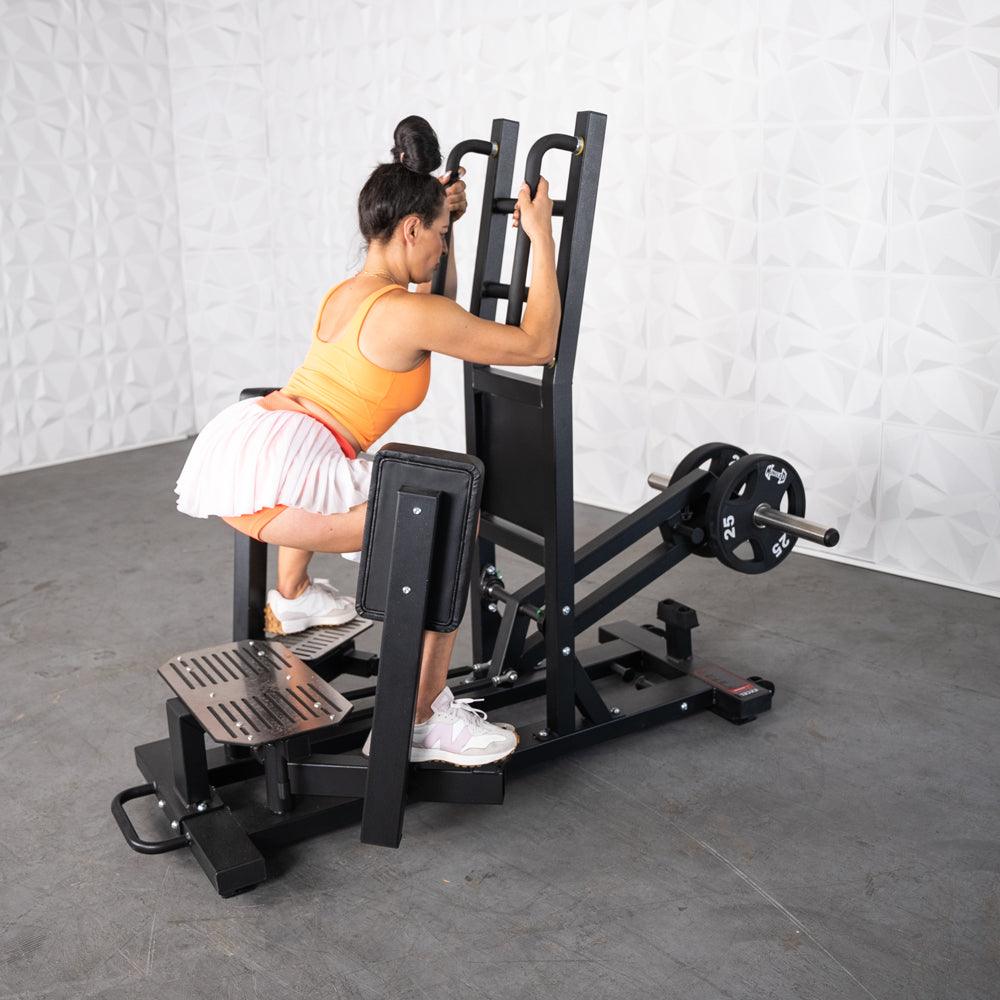 Woman in orange top performing standing hip thrust on a gym machine with weights.
