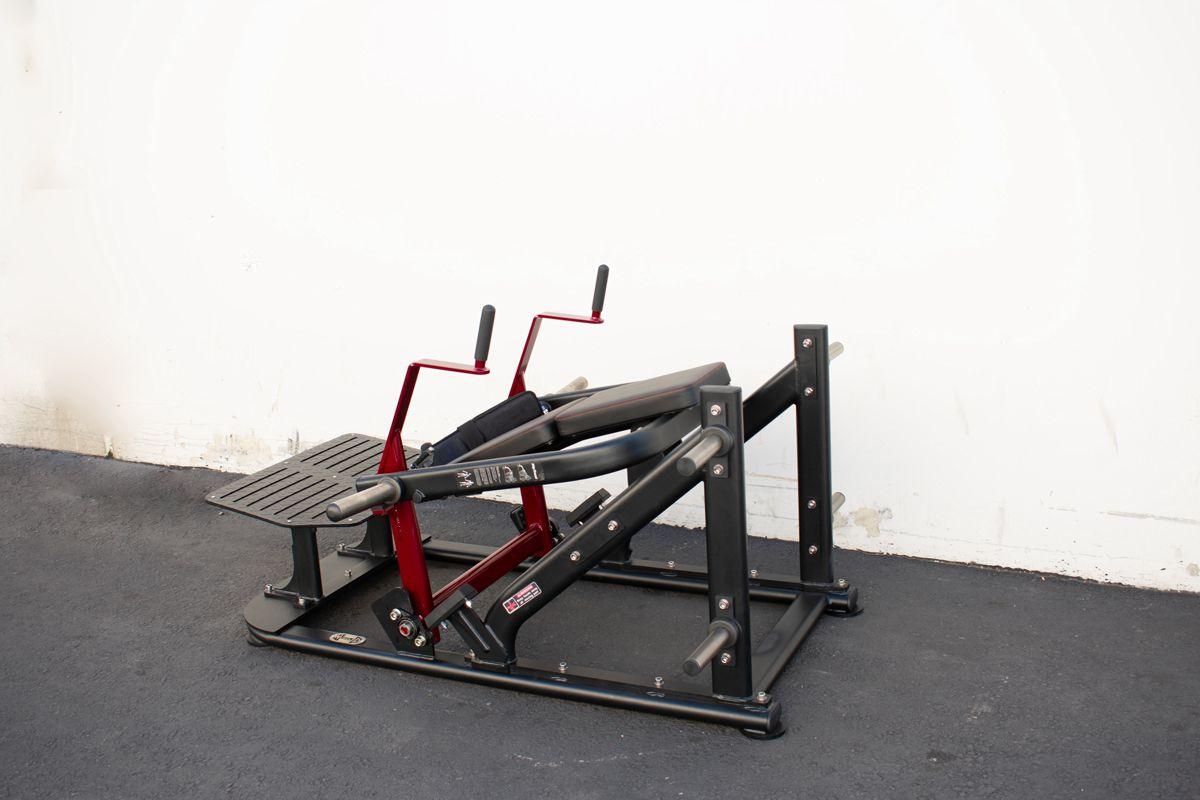 Black and red plate-loaded gym leg press/extension machine on a dark floor.