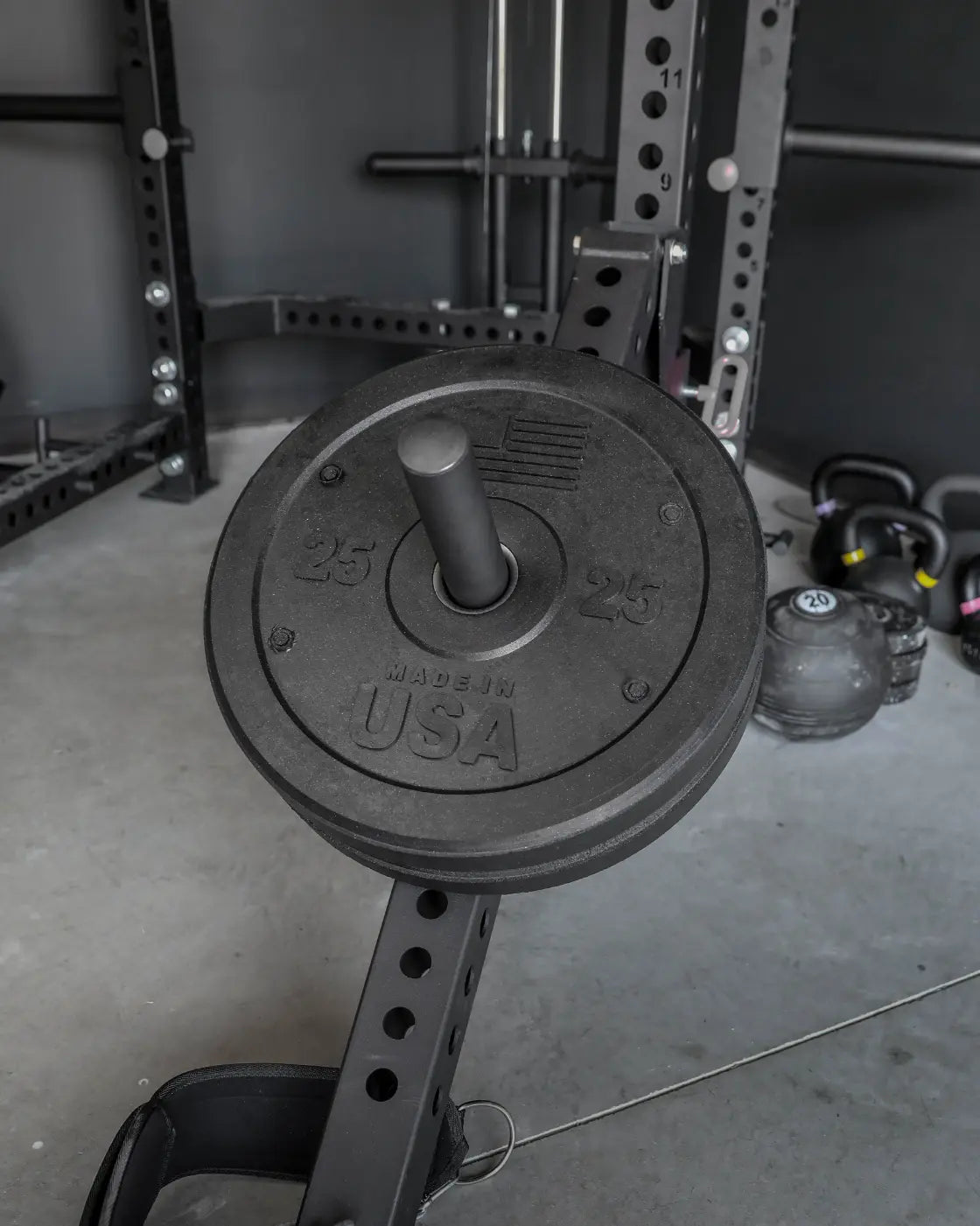 Black bumper plate with 25-pound weight markings and "Made in USA" text, mounted on a barbell in a gym setting with a power rack and kettlebells visible in the background.