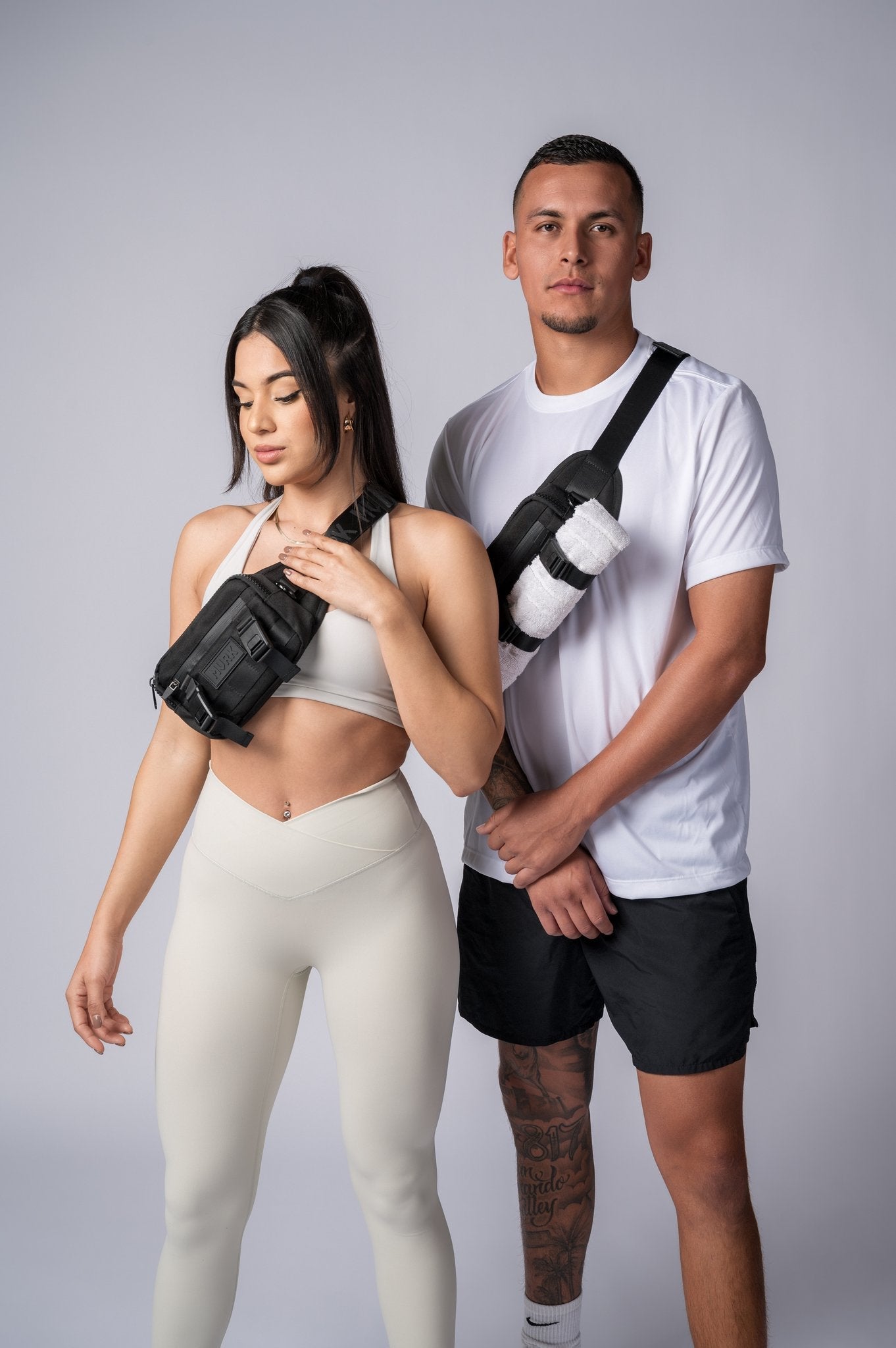 Athletic couple posing in white workout outfits with crossbody bags in studio.