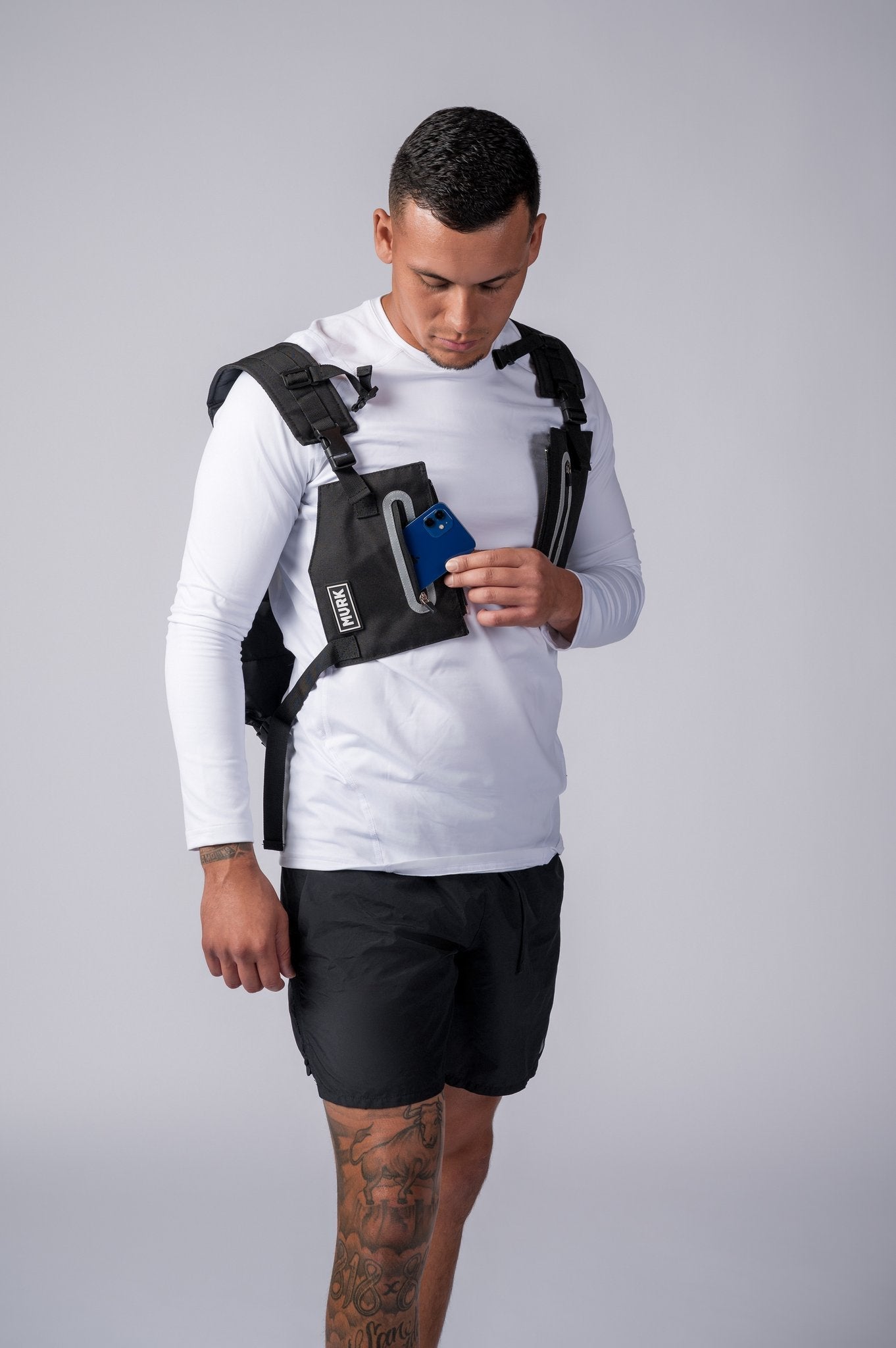 Man in white long-sleeve shirt wearing a tactical vest, holding a blue phone.