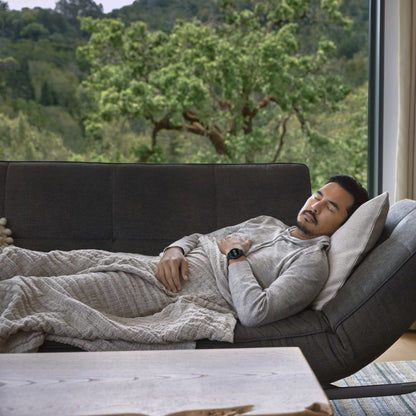 A man resting on a couch, covered with a blanket, wearing a Garmin Vivoactive 5 smartwatch, with a scenic view of greenery outside the window.