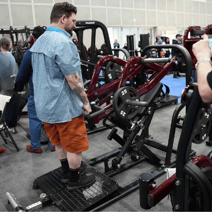 Man using a red leverage squat machine at a fitness expo.