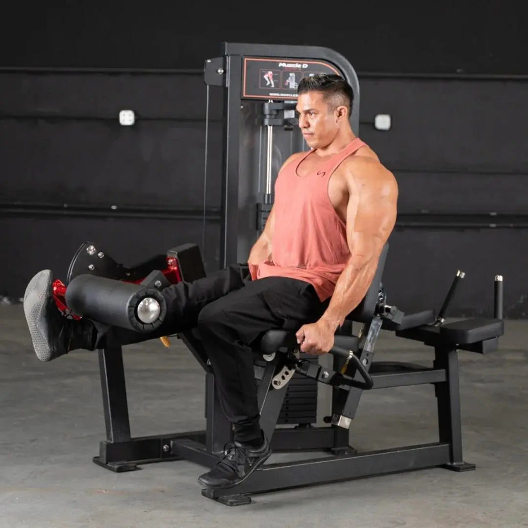 Man performing leg extension on a black Muscle D Fitness leg extension and lying leg curl combo machine in a gym with dark walls.