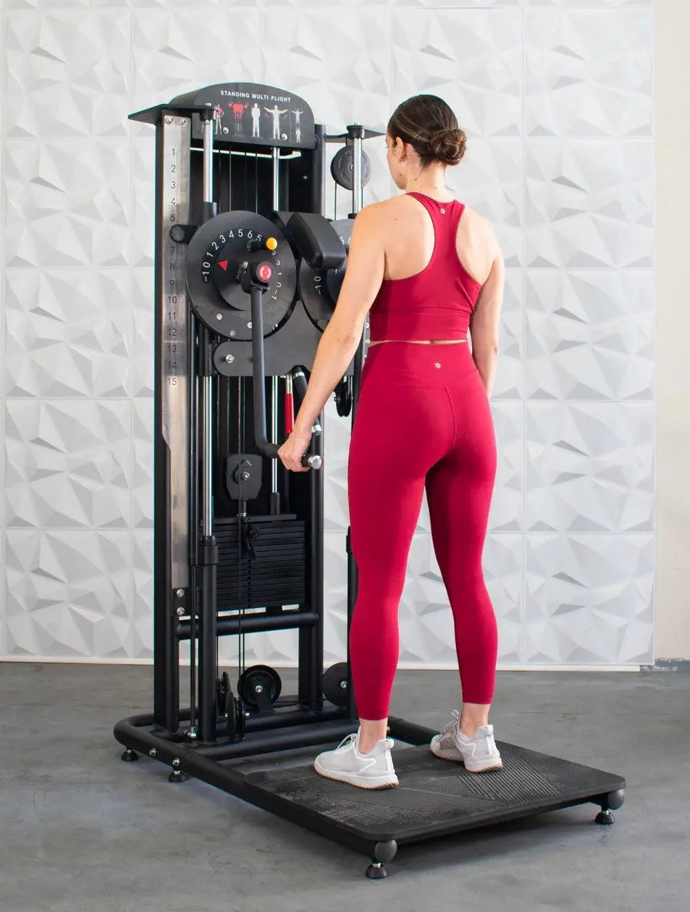 Woman in red workout attire using a black standing multi-flight trainer machine with weight stack and exercise instruction graphics on white textured wall background.