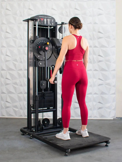 Woman in red workout attire using a black standing multi-flight trainer machine with weight stack and exercise instruction graphics on white textured wall background.