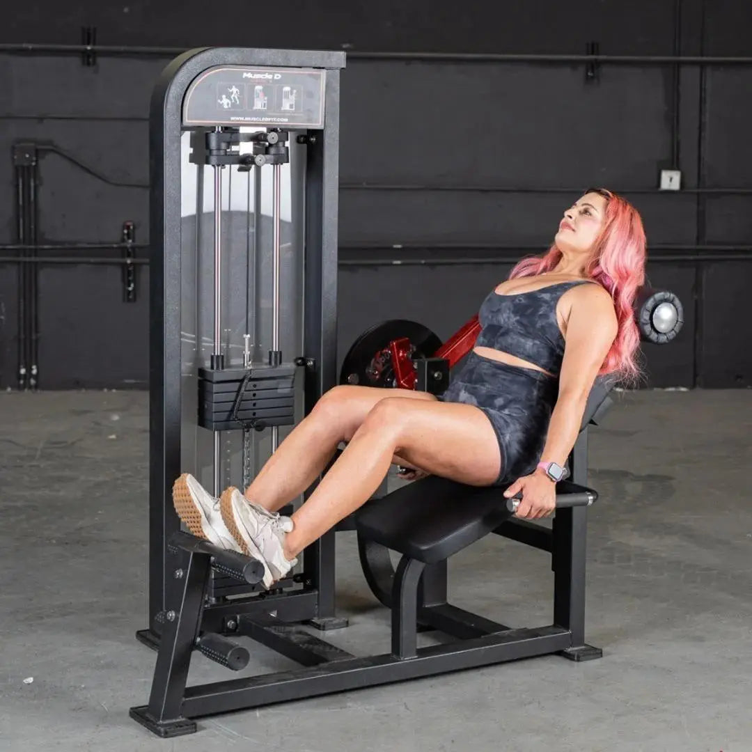 Woman performing a leg extension exercise on a black selectorized leg extension and ab machine in a gym with dark walls.