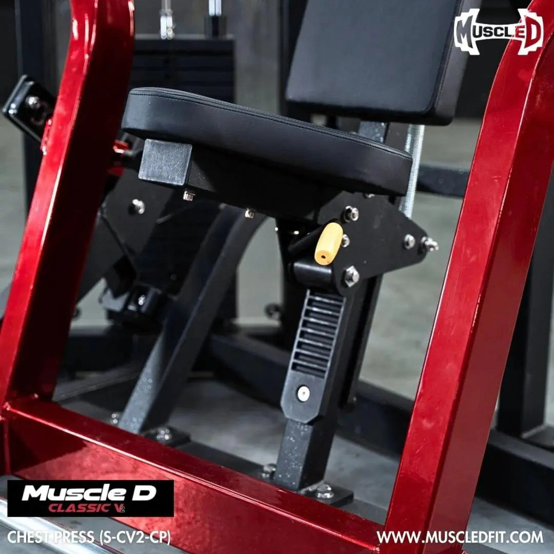 Close-up of chest press machine with red frame, black padded seat and backrest, yellow grip handles, and metal adjustment mechanisms in a gym setting.