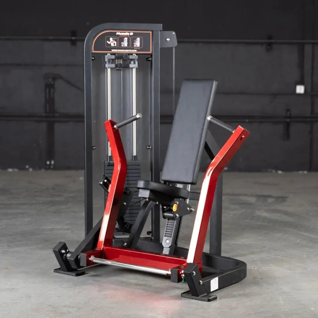 Commercial chest press machine with red and black frame, dual vertical cylinders, gray padded seat and backrest, and instruction panel at top on concrete gym floor.