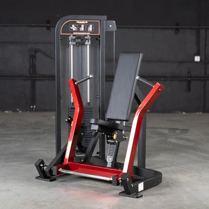 Commercial chest press machine with red and black frame, dual vertical cylinders, gray padded seat and backrest, and instruction panel at top on concrete gym floor.