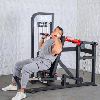 Man performing a chest and shoulder press on a black selectorized machine with red arm handles and cable pulley system against a textured white wall.