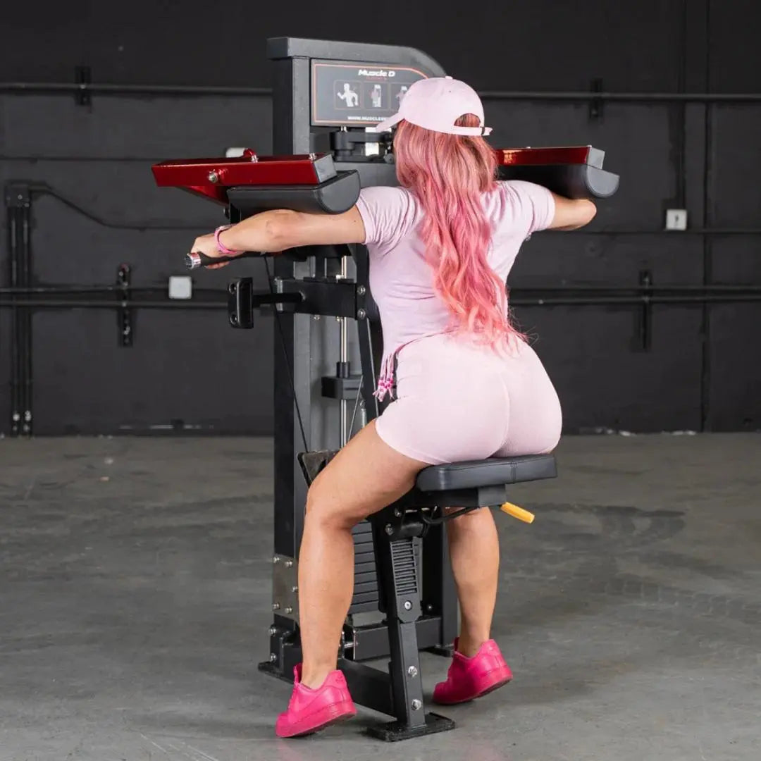 Woman performing a lateral raise on a Muscle D selectorized machine with red handles and black frame in a gym setting.