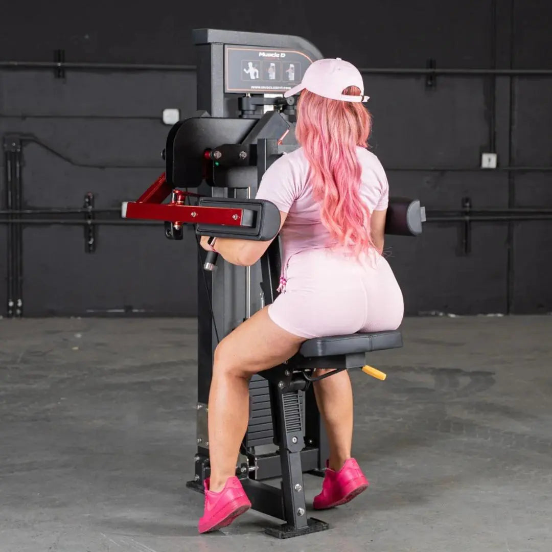 Woman performing a lateral raise exercise on a black and red selectorized machine with instruction display panel in a gym setting.