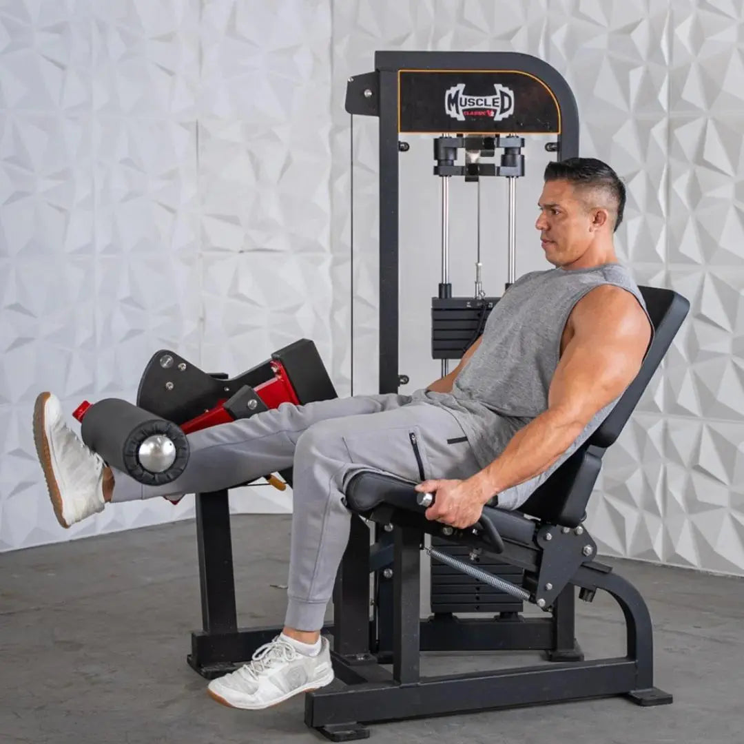 Man performing leg extension on a black Muscle D selectorized leg extension machine with orange trim against a white textured wall.