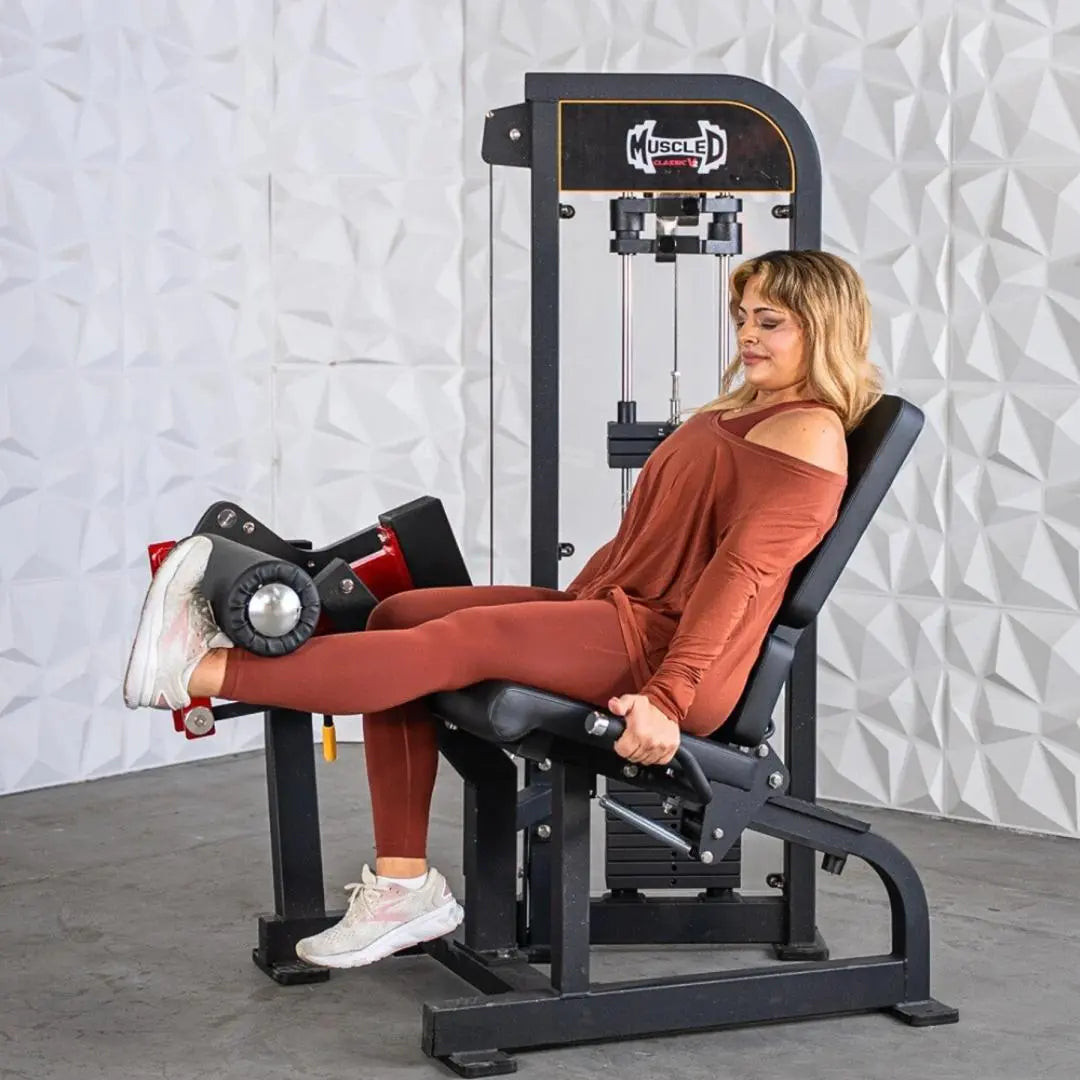 Woman performing a leg extension exercise on a black and white Muscle D selectorized leg extension machine with red weight stack selector and foot pad.