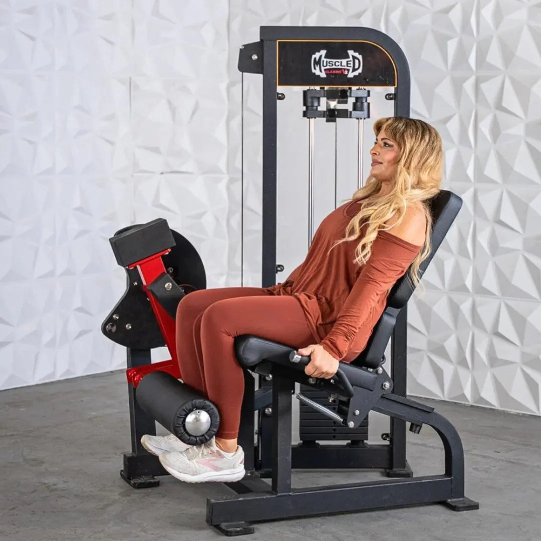 Woman performing leg extension on a black and red selectorized leg extension machine with cable-driven system and padded seat.