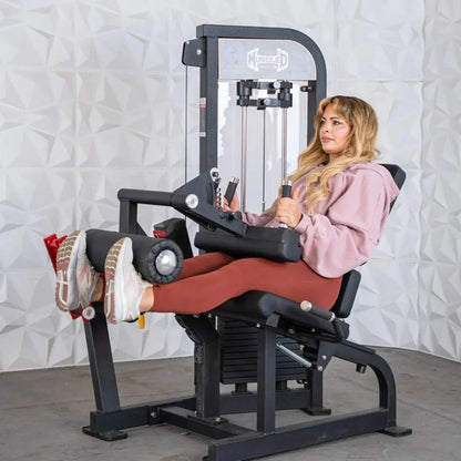 Woman performing a seated leg curl on a black and red Muscle D Fitness leg extension and curl combo machine with a white textured wall backdrop.