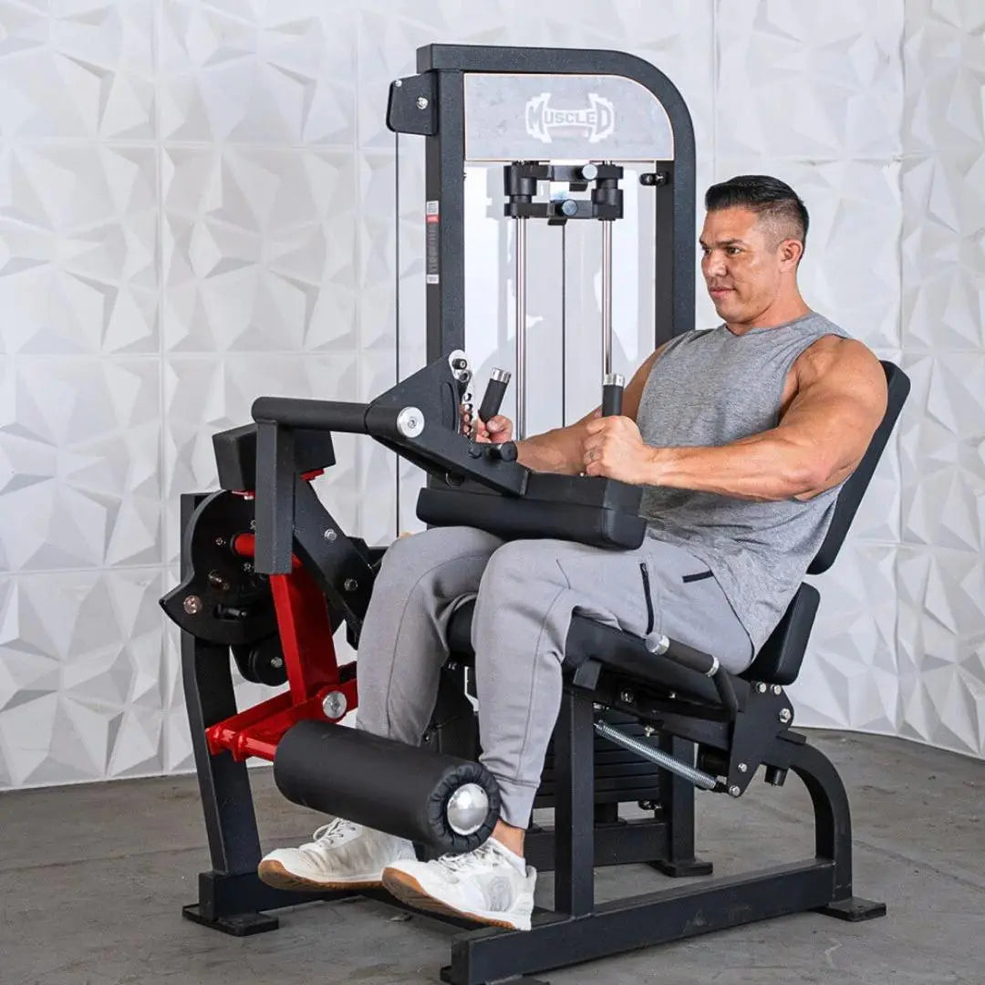 Man performing a seated leg curl on a black and red selectorized leg extension and curl combo machine with white textured wall backdrop.