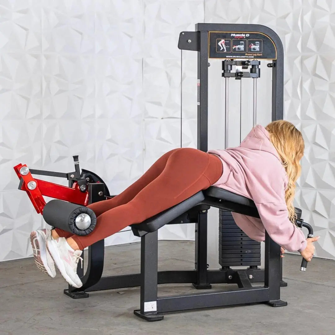 Woman performing prone leg curl on a black Muscle D selectorized leg curl machine with red foot platform and adjustment handles.