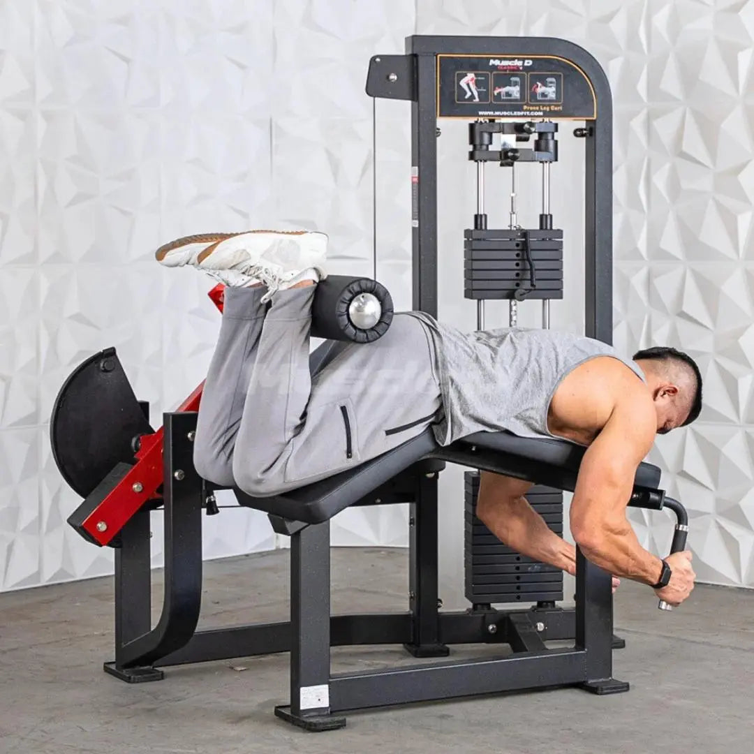 Man performing prone leg curl on a black and red selectorized leg curl machine with stack weights and instruction panel visible.